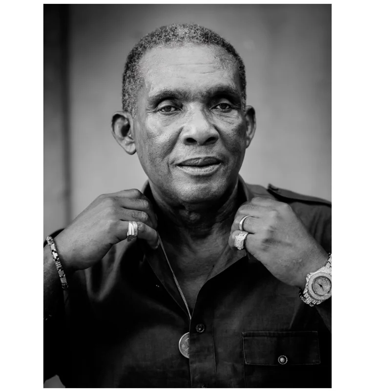 Czarno-biały portret Kena Boothe, legendarnego wokalisty reggae z projektu Reggae Faces, fot. Bartek Muracki. Ken Boothe patrzy się w obiektyw, poprawia kołnierzyc koszuli obiema rękami. Na lewym nadgarstku ma zegarek, a na palcach sygnety.
