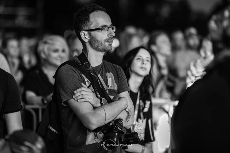 Portret Bartka Murackiego przed sceną, Bartek stoi z założonymi rękami i ma na sobie sprzęt fotograficzny